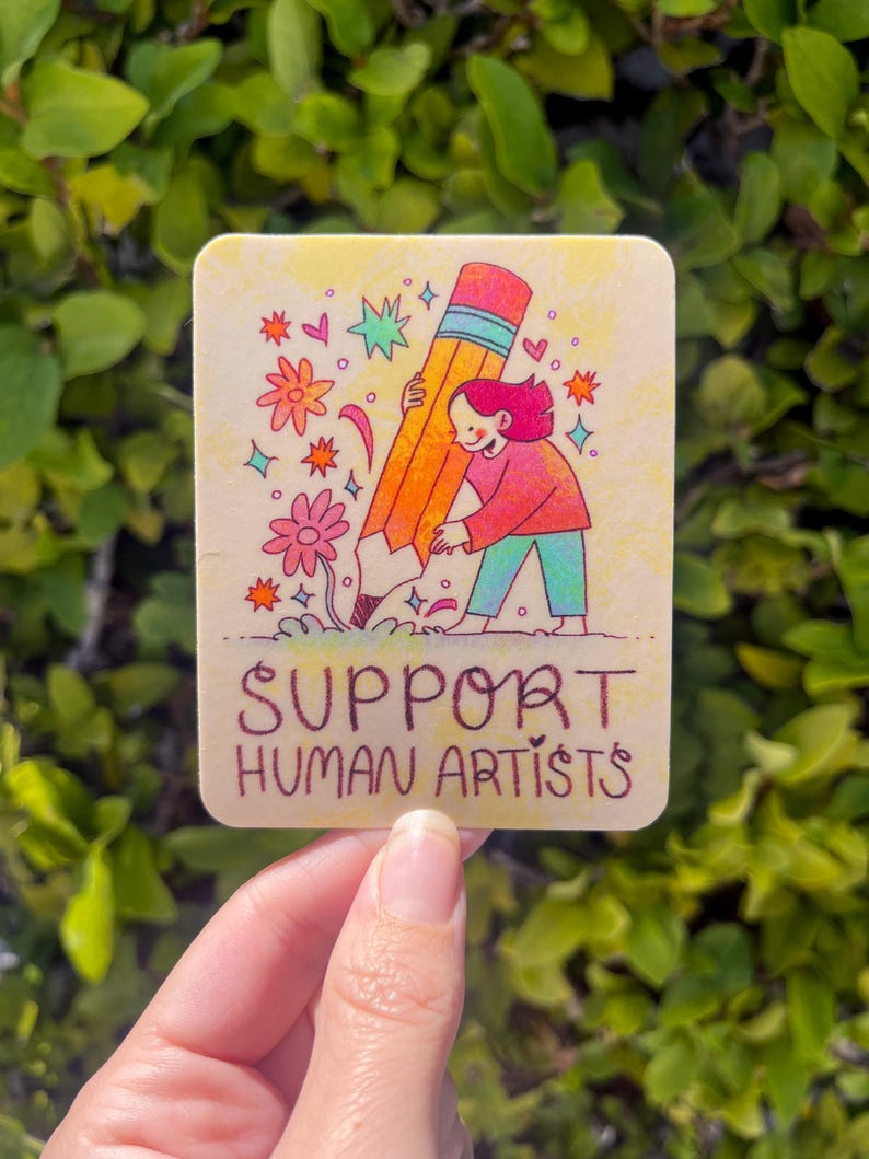 May include: A sticker with a cartoon illustration of a person holding a large pencil with the text "SUPPORT HUMAN ARTISTS" below.