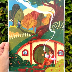 Op de afbeelding: Een kleurrijke illustratie van een hobbit die buiten een ronde deur zit met een pijp in zijn mond. De hobbit draagt een bruin vest en broek. De deur is groen met een messing handvat. Er is een wit hek op de achtergrond en een klein figuur in de verte.