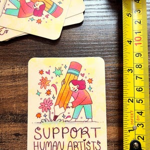May include: A yellow card with a cartoon illustration of a person holding a large pencil. The card has the text "SUPPORT HUMAN ARTISTS" in black.