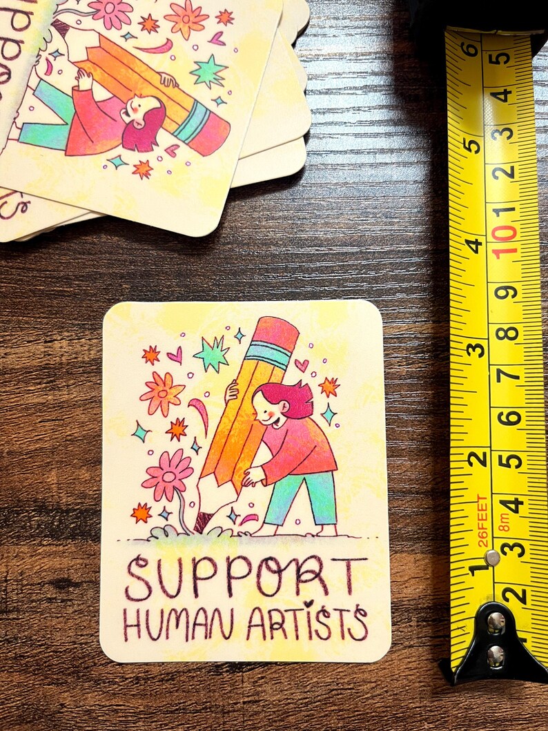 May include: A yellow card with a cartoon illustration of a person holding a large pencil. The card has the text "SUPPORT HUMAN ARTISTS" in black.