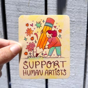 May include: A yellow and orange pencil is being planted in the ground by a person with red hair. The pencil is surrounded by flowers and stars. The text "SUPPORT HUMAN ARTISTS" is written below the image.