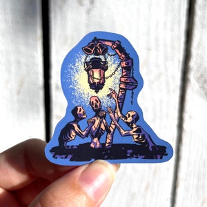 Pu&ograve; includere: Un adesivo con un'illustrazione blu e viola di tre figure scheletriche che raggiungono una lanterna incandescente appesa a un palo di legno.