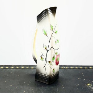 Könnte beinhalten: Eine weiße Keramikvase mit schwarzem und goldenem Rand. Die Vase hat eine einzigartige Form und zeigt ein gemaltes Design eines Zweigs mit grünen Blättern und einem roten Apfel.