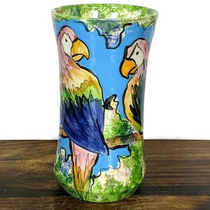 May include: A ceramic vase with a blue background and two parrots painted on it. The parrots are yellow, green, blue, and pink. The vase has a green, white, and blue speckled glaze.