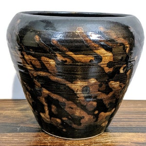 May include: A brown and black ceramic planter with a unique, abstract pattern. The planter has a wide, round opening and a slightly tapered body.