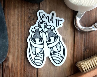 May include: A sticker featuring the phrase "Send It" and a drawing of climbing shoes. The image includes climbing equipment such as tape, a carabiner, a brush, and climbing shoes, all arranged on a wooden background.