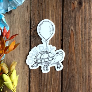 May include: A black and white sticker of a turtle holding a balloon with the word "ONE DAY" written on the shell.
