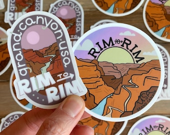 Op de afbeelding: Twee ronde stickers met een panoramisch uitzicht op de Grand Canyon. De stickers zijn wit met een paars, oranje en blauw ontwerp. De tekst op de stickers luidt "Rim to Rim" en "Grand Canyon USA".