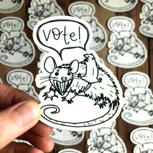 May include: A black and white sticker featuring a cartoon rat with a smaller rat riding on its back. The larger rat has a toothy grin and the sticker has the word "Vote!" in a speech bubble above the rats.