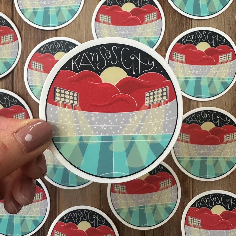 May include: Round sticker with a Kansas City stadium design. The sticker features a red stadium, a yellow sun, and the words "Kansas City" in a black, stylized font. The background has blue and gray stripes.