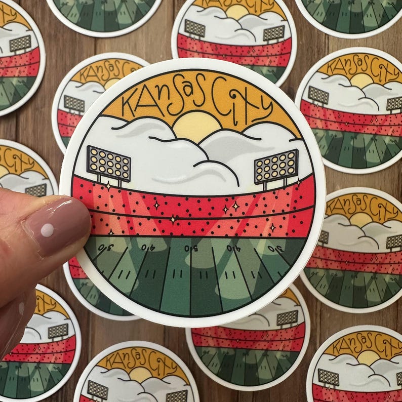 May include: A round sticker featuring a stadium scene with the text "Kansas City". The design includes a green field, red seating, and a yellow sun behind clouds. Several stickers are visible.