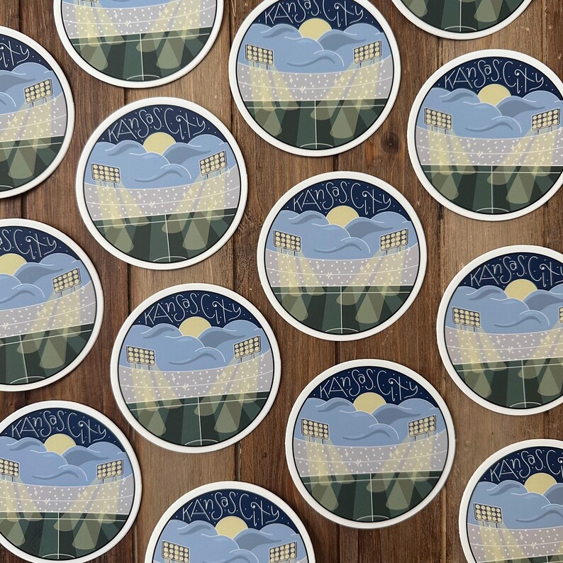 May include: Circular sticker featuring a stadium scene with the text "Kansas City". The design includes a yellow sun, blue clouds, stadium lights, and a green field. The stickers are arranged on a wooden surface.
