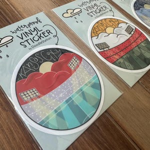 May include: Waterproof vinyl stickers featuring a Kansas City design. The stickers have a circular shape with a white border. The design includes a red cloud, sun, and the text "Kansas City". The stickers are by DoodleStation.