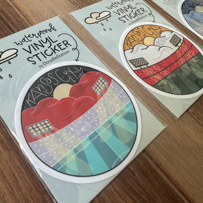 May include: Waterproof vinyl stickers featuring a Kansas City design. The stickers have a circular shape with a white border. The design includes a red cloud, sun, and the text "Kansas City". The stickers are by DoodleStation.
