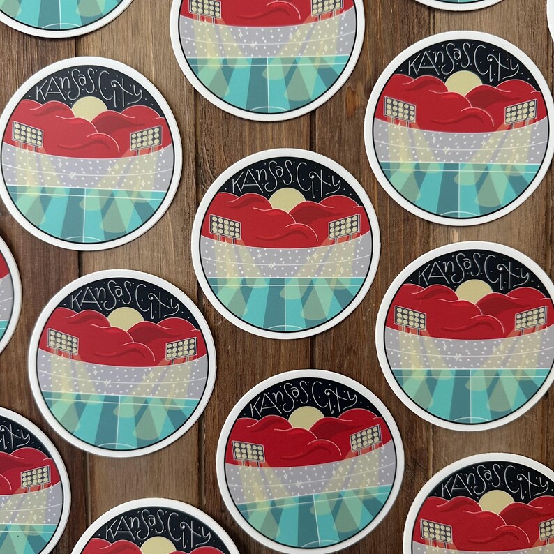 May include: Round stickers with a Kansas City design. The design features red stadium seating, a yellow sun, and the words "Kansas City" in a stylized font. The background includes blue and gray stripes.