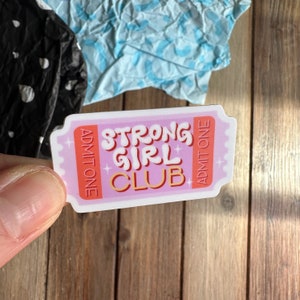 May include: Pink and white sticker shaped like a ticket with the text "Strong Girl Club" in orange and red lettering. The sticker has a gold star design and the words "Admit One" on either side.