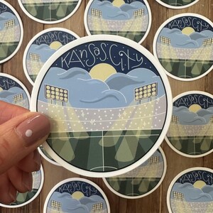 May include: Round sticker featuring a stadium scene with the text "Kansas City" in a stylized font. The design includes a yellow sun, blue clouds, and stadium lights. The sticker has a white border and is surrounded by other similar stickers.