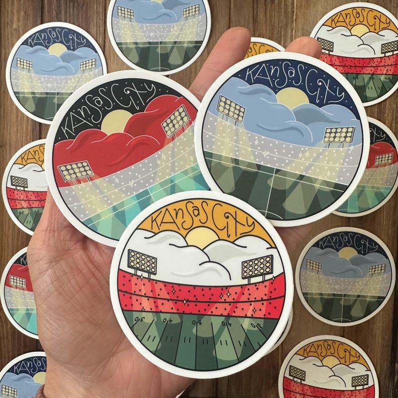 May include: Round stickers featuring a stadium design with the text "Kansas City". The stickers have a colorful illustration of a stadium with a football field, stadium lights, and clouds. The stickers are white with a black border.