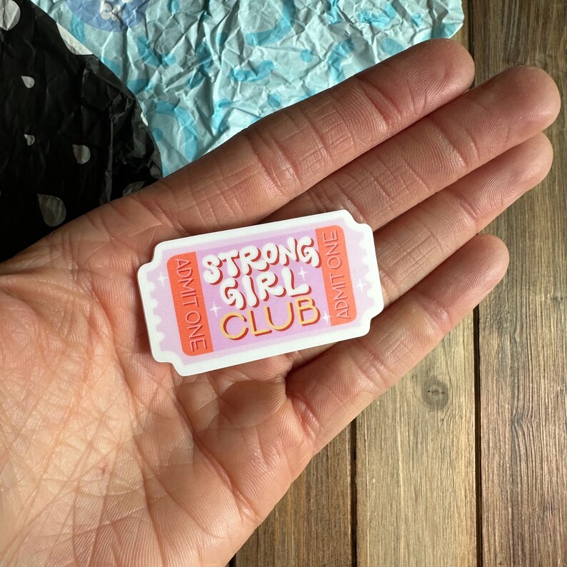 May include: Pink and white ticket-shaped sticker with the text "Strong Girl Club" in orange and pink lettering. The sticker has the words "Admit One" on both sides.