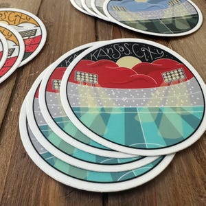 May include: A stack of round coasters with a stadium design. The coasters feature the text "Kansas City" in a stylized font, with a red and yellow stadium graphic and a teal and green field.