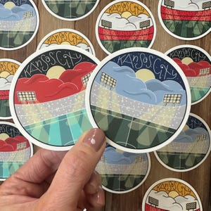 May include: A collection of round stickers featuring a stadium design with the text "Kansas City". The stickers have a white border and depict a stadium with a sun, clouds, and stadium lights. The stadium is colored in red, blue, and green.