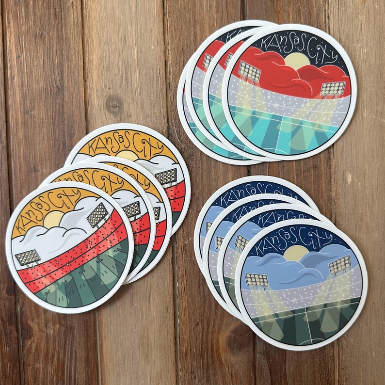 May include: A collection of round stickers featuring a stadium scene with the text "Kansas City." The stickers have a colorful design with a sunset, clouds, and stadium lights. The stickers are arranged on a wooden surface.