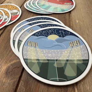 May include: A stack of round coasters with a white border. Each coaster features a stadium scene with the text "Kansas City" in a stylized font. The design includes a blue sky, stadium lights, and a sun.