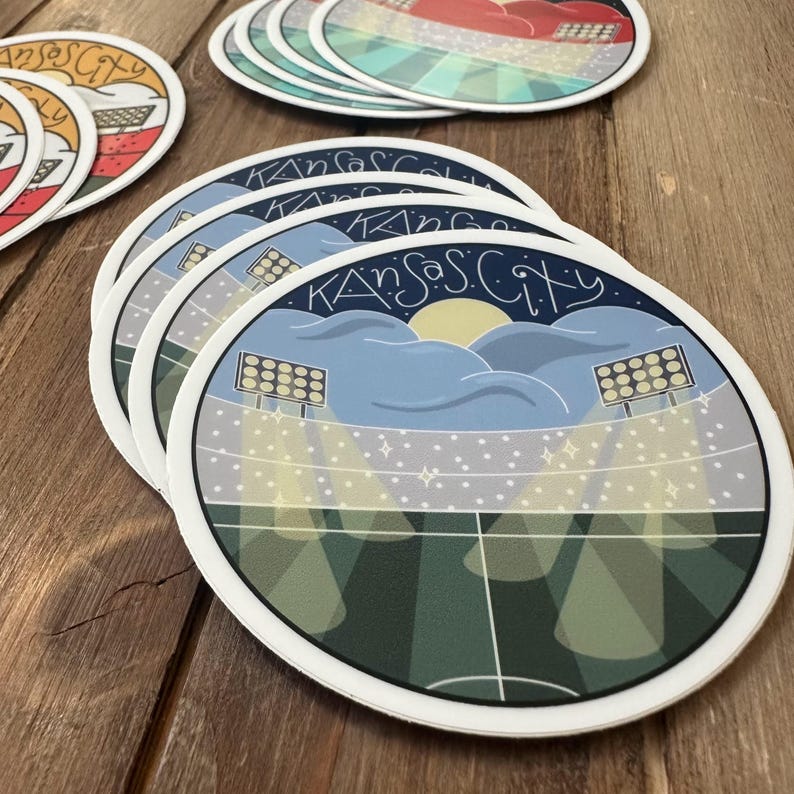 May include: A stack of round coasters with a white border. Each coaster features a stadium scene with the text "Kansas City" in a stylized font. The design includes a blue sky, stadium lights, and a sun.