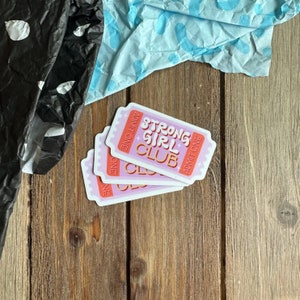 May include: Three pink and white ticket-shaped stickers with the text "Strong Girl Club" in orange and white lettering. The text "Admit One" is printed on each sticker.