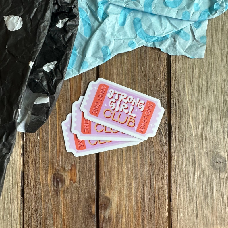 May include: Three pink and white ticket-shaped stickers with the text "Strong Girl Club" in orange and white lettering. The text "Admit One" is printed on each sticker.