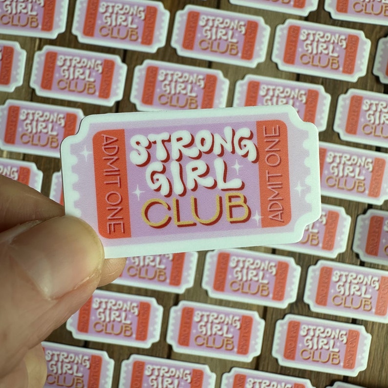 May include: A pink and orange ticket sticker with the text "Strong Girl Club" and "Admit One" printed on it. The sticker is being held by a hand.