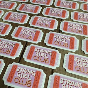 May include: Multiple pink and orange stickers with the text "Strong Girl Club" in a retro font. The stickers are shaped like tickets and have the words "Admit One" on them.