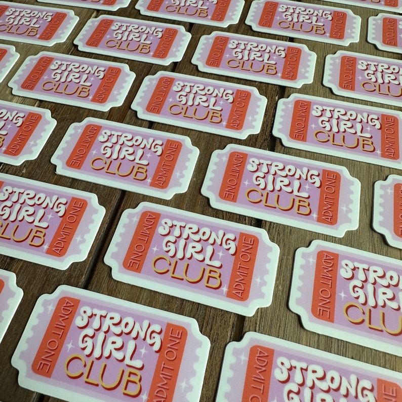 May include: Multiple pink and orange stickers with the text "Strong Girl Club" in a retro font. The stickers are shaped like tickets and have the words "Admit One" on them.