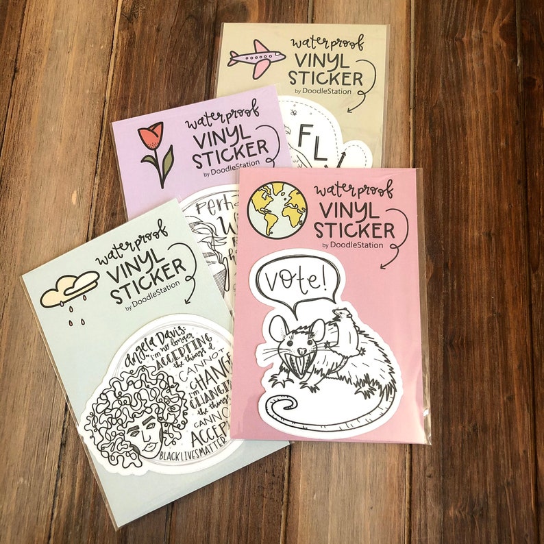 May include: Four waterproof vinyl stickers with different designs. The stickers are packaged in clear plastic sleeves. The designs include a cloud with raindrops, a globe, a rat with a speech bubble that says "vote!" and a portrait of Angela Davis with the text "Angela Davis I'm no longer accepting the things I cannot change" and "Black Lives Matter".