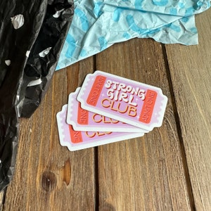 May include: Three pink and white ticket-shaped stickers with the text "Strong Girl Club" in orange and white letters. The stickers are stacked on top of each other and have the words "Admit One" on each side.