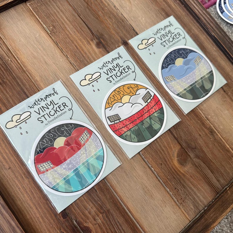 May include: Three waterproof vinyl stickers featuring a circular design with the text "Kansas City" and a landscape illustration. Each sticker has a different color scheme, including red, green, blue, and yellow. The stickers are packaged on light blue backing cards.