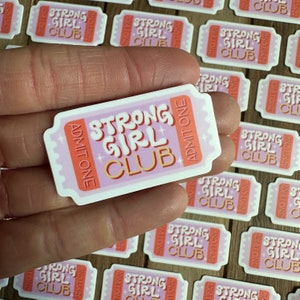 May include: Pink and orange ticket-shaped sticker with the text "Strong Girl Club" and "Admit One" on it.