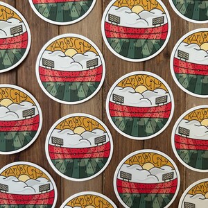 May include: Circular stickers with a stadium design. The stickers feature a red and green field, white clouds, a yellow sun, and the text "Kansas City" in a circular design. The stickers are on a wooden surface.