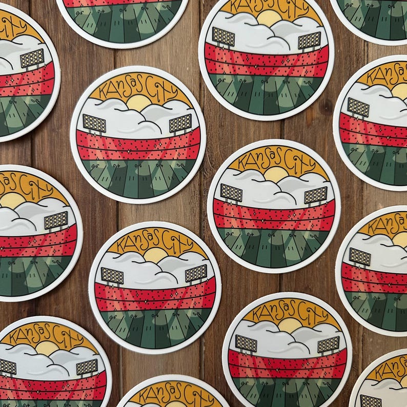 May include: Circular stickers with a stadium design. The stickers feature a red and green field, white clouds, a yellow sun, and the text "Kansas City" in a circular design. The stickers are on a wooden surface.