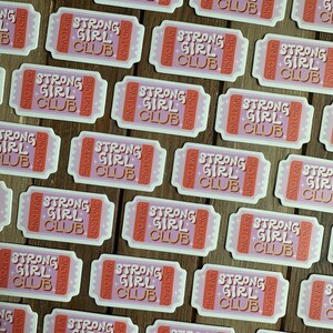May include: Multiple pink and white ticket-shaped stickers with the text "Strong Girl Club" in gold lettering. The stickers have a retro design with a pink background and white lettering.