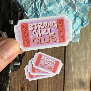 May include: Four pink and white ticket-shaped stickers with the text "Strong Girl Club" in pink and orange letters. The stickers have a gold star design and the words "Admit One" on each side.