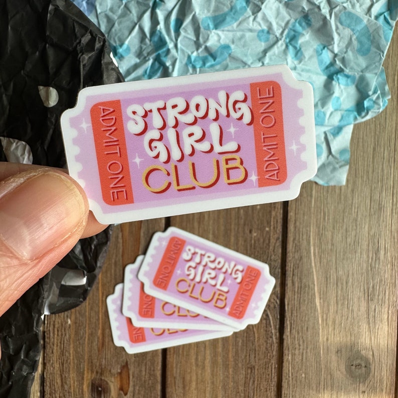 May include: Four pink and white ticket-shaped stickers with the text "Strong Girl Club" in pink and orange letters. The stickers have a gold star design and the words "Admit One" on each side.