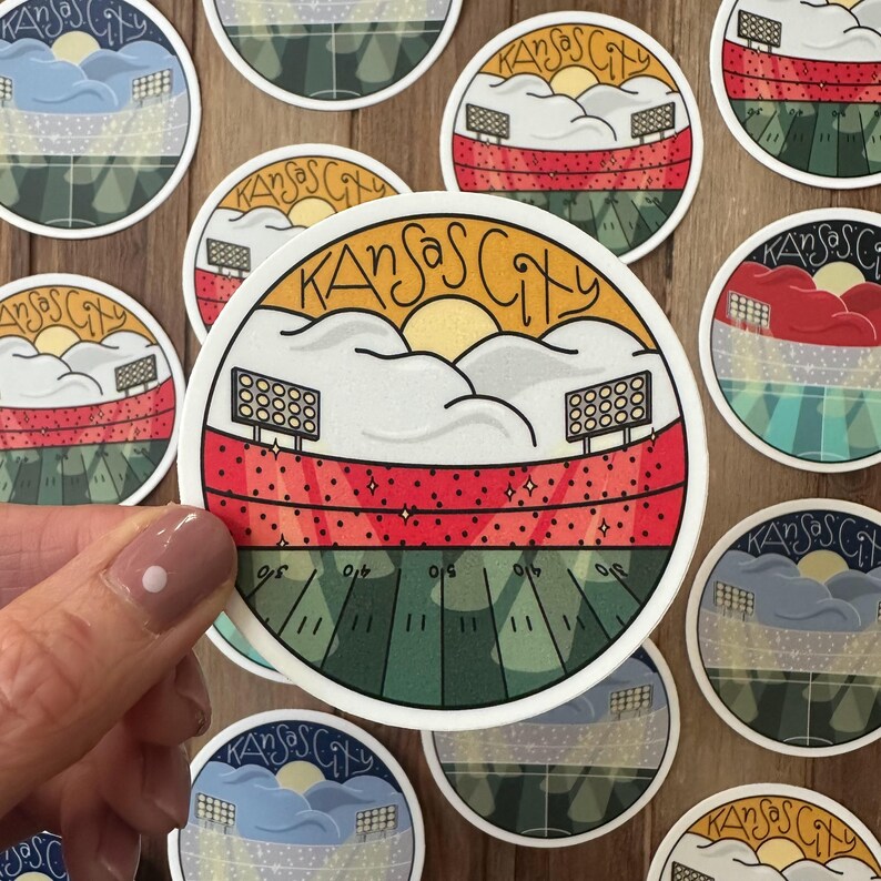 May include: Round sticker featuring a stadium scene with the text "Kansas City". The design includes a football field, stadium lights, and a sunset. The sticker has a white border and is on a wooden surface.