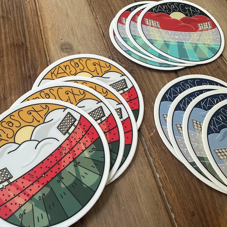 May include: A collection of round stickers featuring a stadium design with the text "Kansas City". The stickers have a white border and depict a football field with red, green, and blue colors, and a sun and clouds.