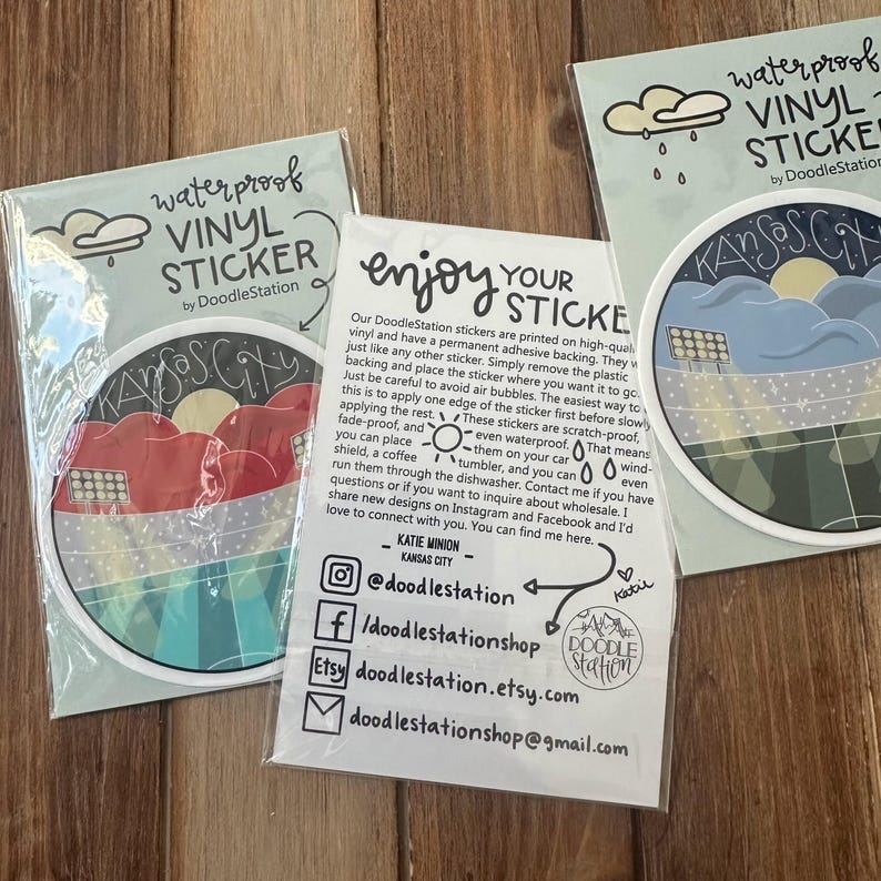 May include: Waterproof vinyl stickers by DoodleStation featuring a Kansas City design. The stickers are round with a colorful illustration of a cityscape, clouds, and a sun. The packaging includes instructions and social media information.