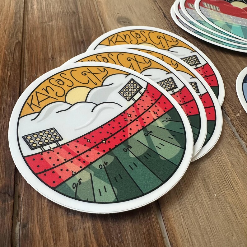 May include: Round stickers featuring a stadium design with the text "Kansas City". The design includes a red and green field, stadium lights, and a yellow sun. The stickers are white with a glossy finish.