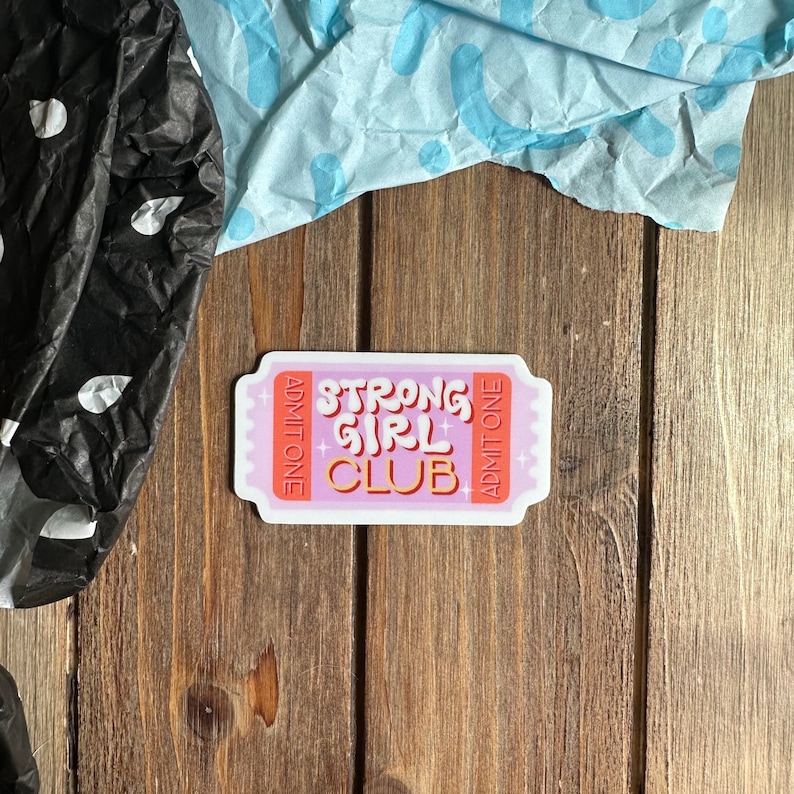 May include: Pink and orange sticker with the text "Strong Girl Club" and "Admit One" on either side.