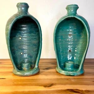 May include: Two teal ceramic candle holders with a bottle-like shape and a large opening. The holders have a small base and a cylindrical candle cup. The interior has a textured surface and a few small holes.