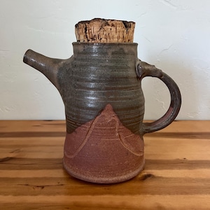 May include: Handmade ceramic teapot with a cork stopper. The teapot is primarily gray with a reddish-brown base and handle. The design includes a mountain-like relief on the lower portion of the pot. The spout and handle are dark brown.