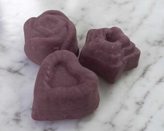 May include: Three purple soap bars in different shapes: a rose, a heart, and a star. The soap bars are on a white marble surface.